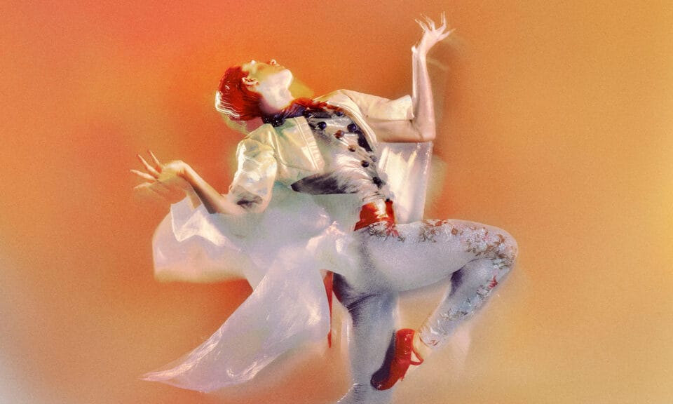 A pale dancer with short red hair dressed like a matador in a white, floral-embroidered outfit with red slippers throws their head back and lifts a leg against a sunset-colored background.