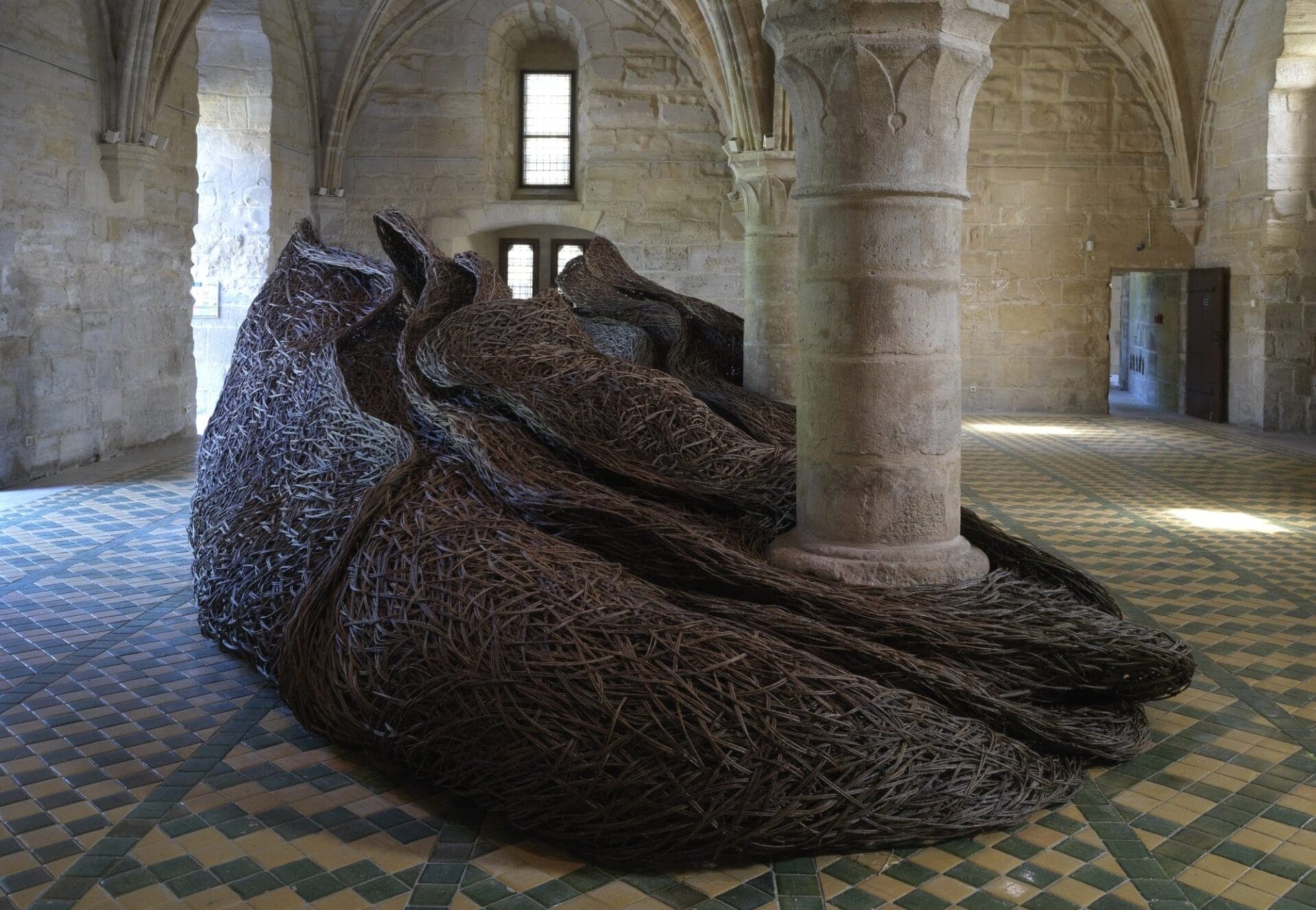 an organic sculptural form made from willow and tree branches wraps around a column in the room of an abbey