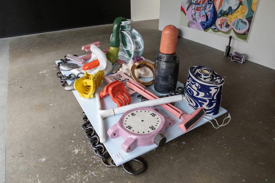 displayed in a gallery atop a platform is an assortment of large-scale ceramic sculptures of everyday objects such as lipstick, claw clips, a clock, perfume bottles, flowers, razors, and an IUD.