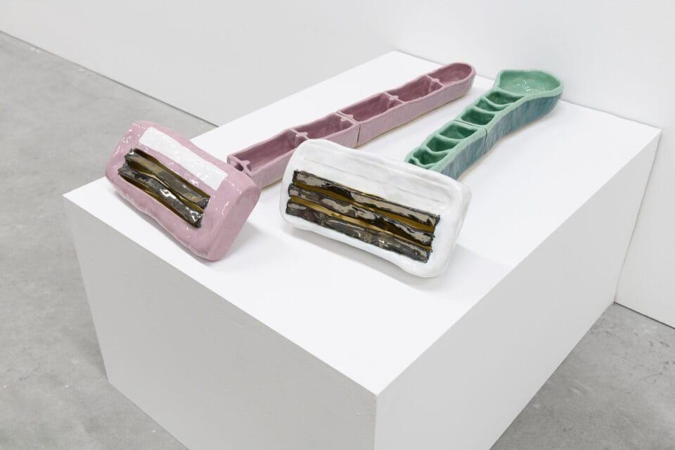 two large scale ceramic sculptures of disposable razors. they sit on a white pedestal in a gallery