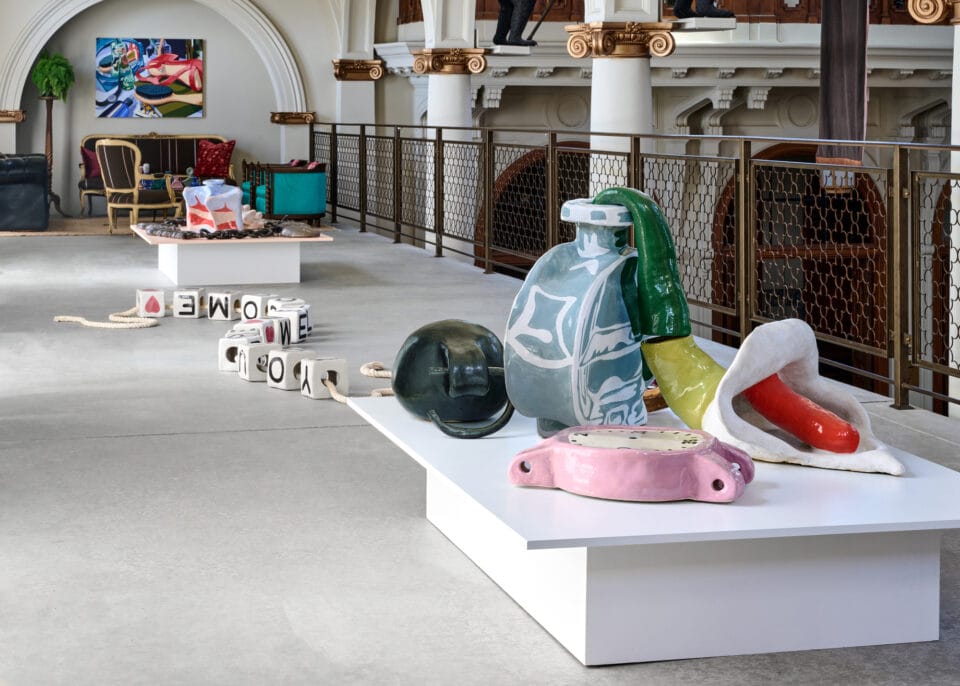 an installation photo of ceramic sculptures placed atop platforms on the floor of a gallery space. the assemblage of sculptures include everyday objects such as clocks, flowers, bottles, and jewelry.