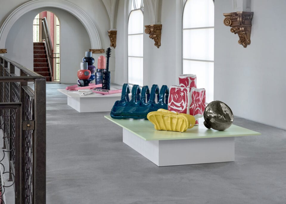 an installation photo of ceramic sculptures placed atop platforms on the floor of a gallery space. the assemblage of sculptures include everyday objects such as makeup, razors, claw clips, ornate perfume bottles, and an IUD