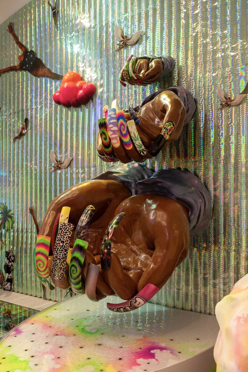 an installation view of a maximalist exhibition featuring numerous collaged photographs, reflective surfaces, and prismatic colors, focused on a stack of hands emerging from the walls, showing highly decorated fingernails