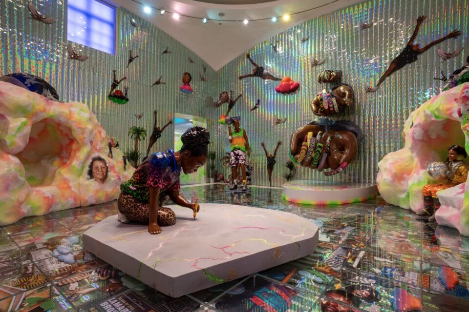 an installation view of a maximalist exhibition by artist Lauren Halsey, featuring numerous collaged photographs, reflective surfaces, Black figures, patterns, and prismatic colors