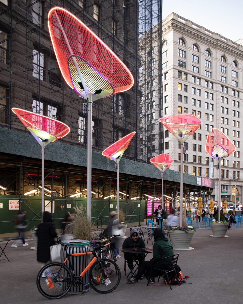 an evening view with people standing and seated around on the sidewalk beneath a series of tulip-like public artworks installed outside of the Flatiron building in New York City