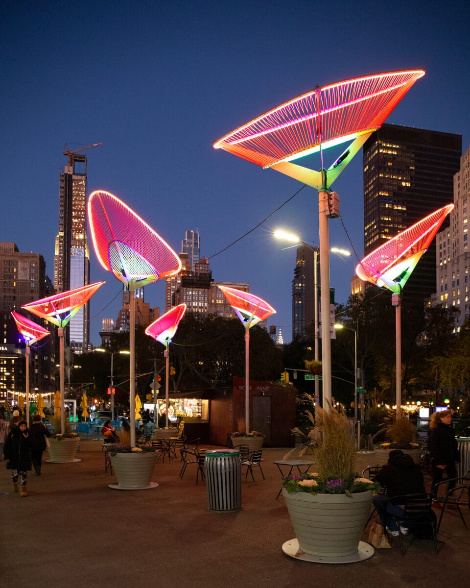 a nighttime view with people standing and seated around on the sidewalk beneath a series of tulip-like public artworks installed outside of the Flatiron building in New York City