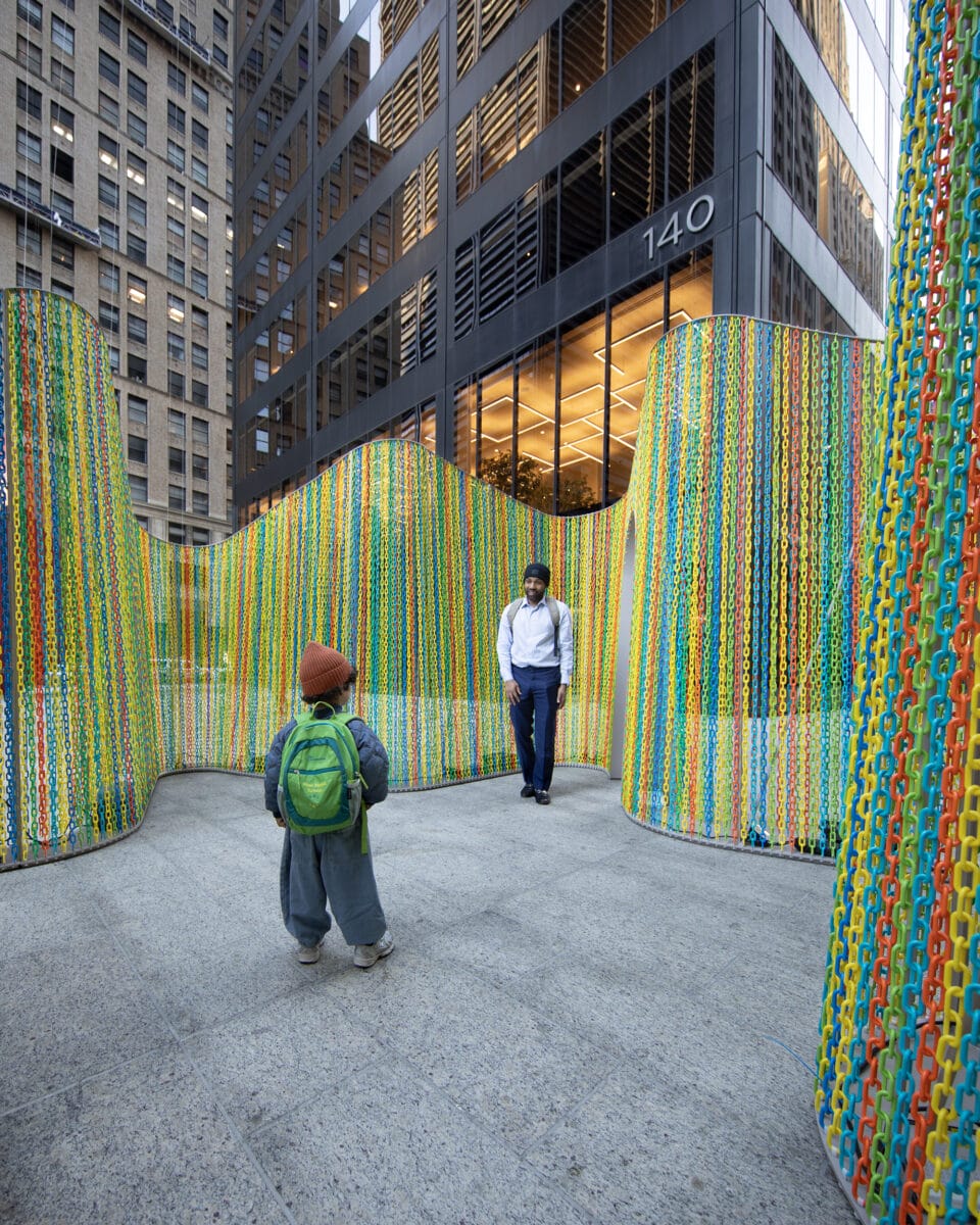 an outdoor installation of undulating, permeable walls with some doorways in mostly bright green and orange, with people standing in the center