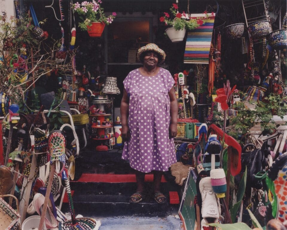 a Black woman in a pink and white polka dot dress and hat standing on the front step of her home with countless found objects sculptures surrounding her