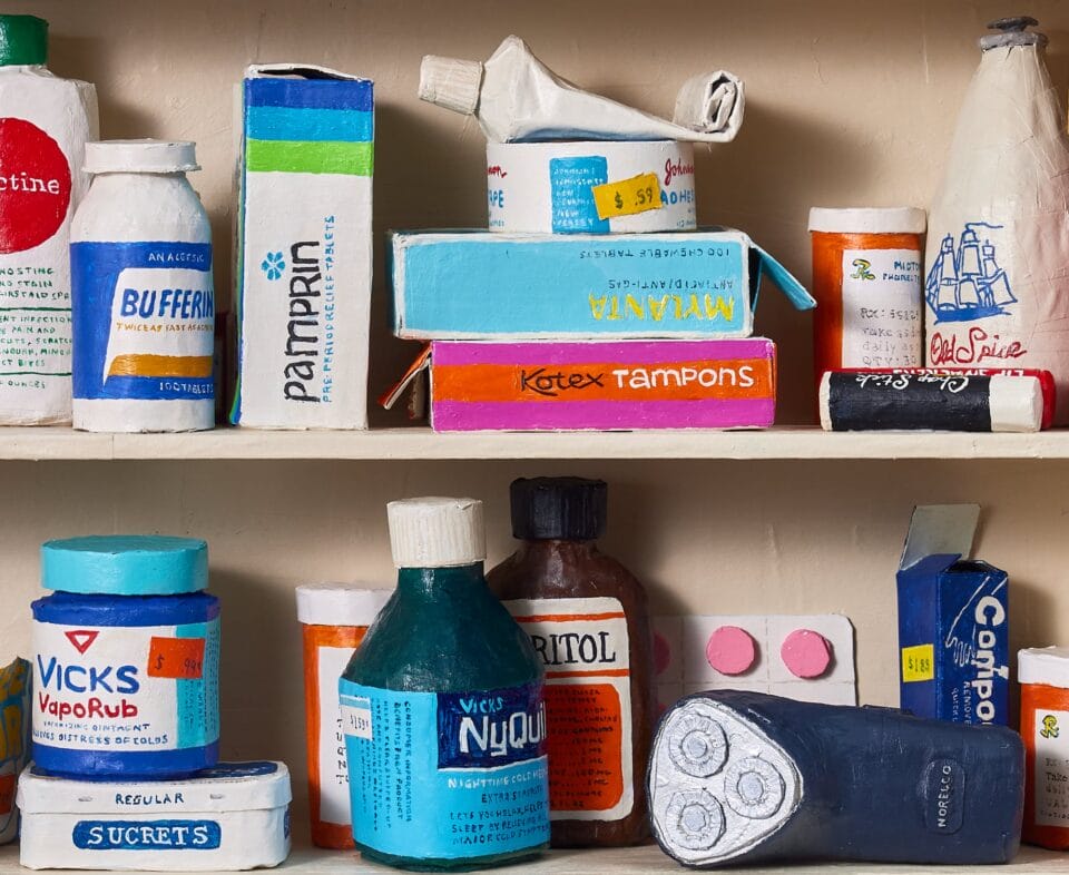 a detail image of a paper mache sculpture of a medicine cabinet filled with over the counter medicines, prescriptions, and other bathroom goods