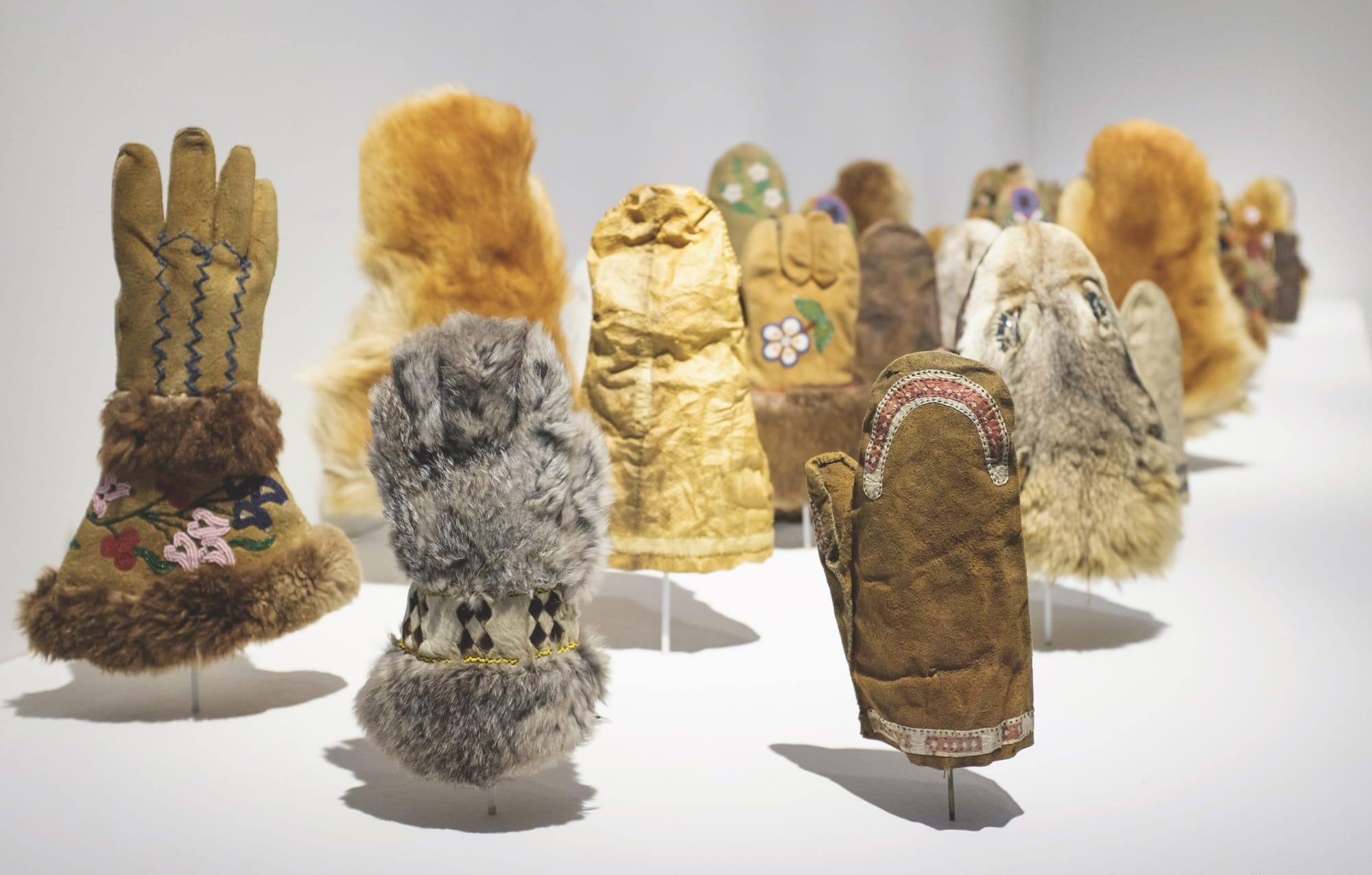 a detail of an installation of various handmade fur and seal skin mittens illuminated in a gallery