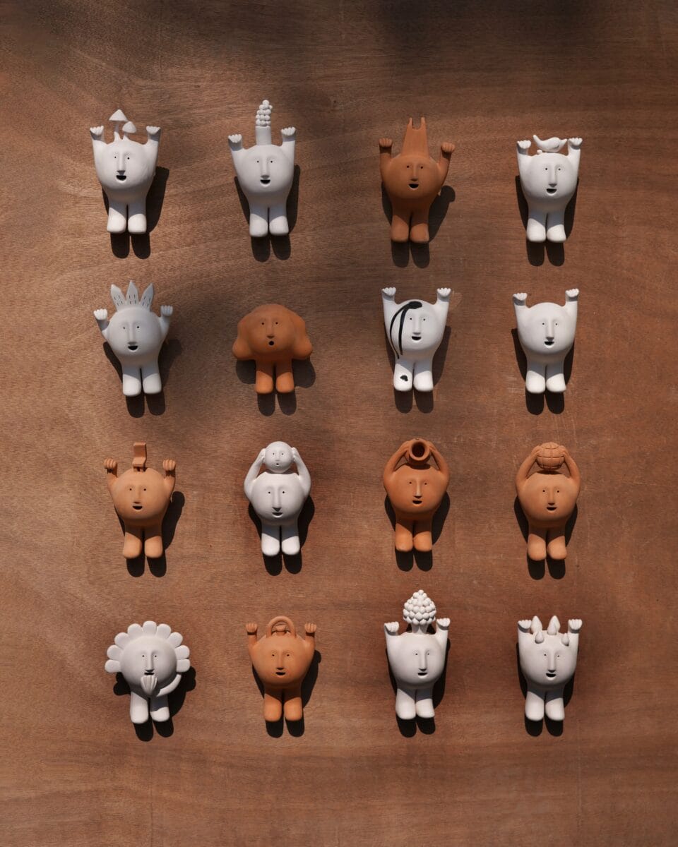 a grid of sixteen small figurines of expressive ceramic faces with limbs, hung on the wall in shades of ivory and terracotta. though they look similar, each dons its own unique feature