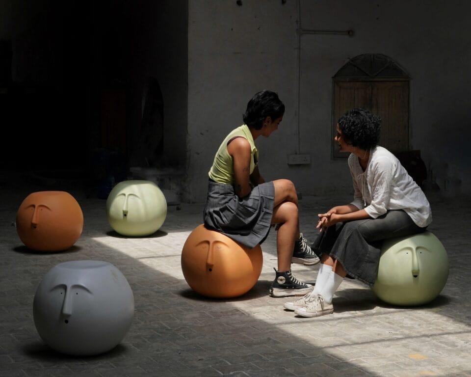 five heads in hues of ivory, sage green, and orange take the form of spherical seats, two individuals rest upon them