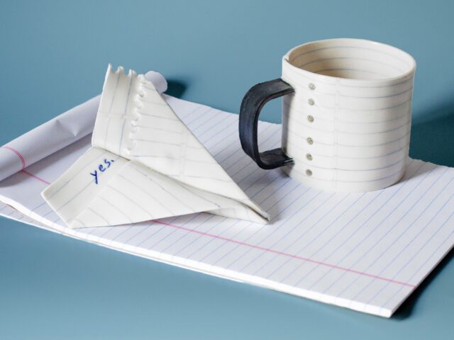 a mug and paper airplane sculpture that appear to made of lined paper atop white notepad