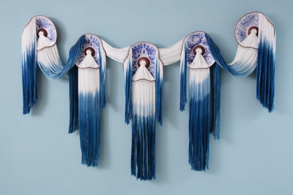 a wall installation of four ceramic plates with blue glaze detail, connected by lengths of blue fiber that drapes between them and emerges from holes in the center of each plate