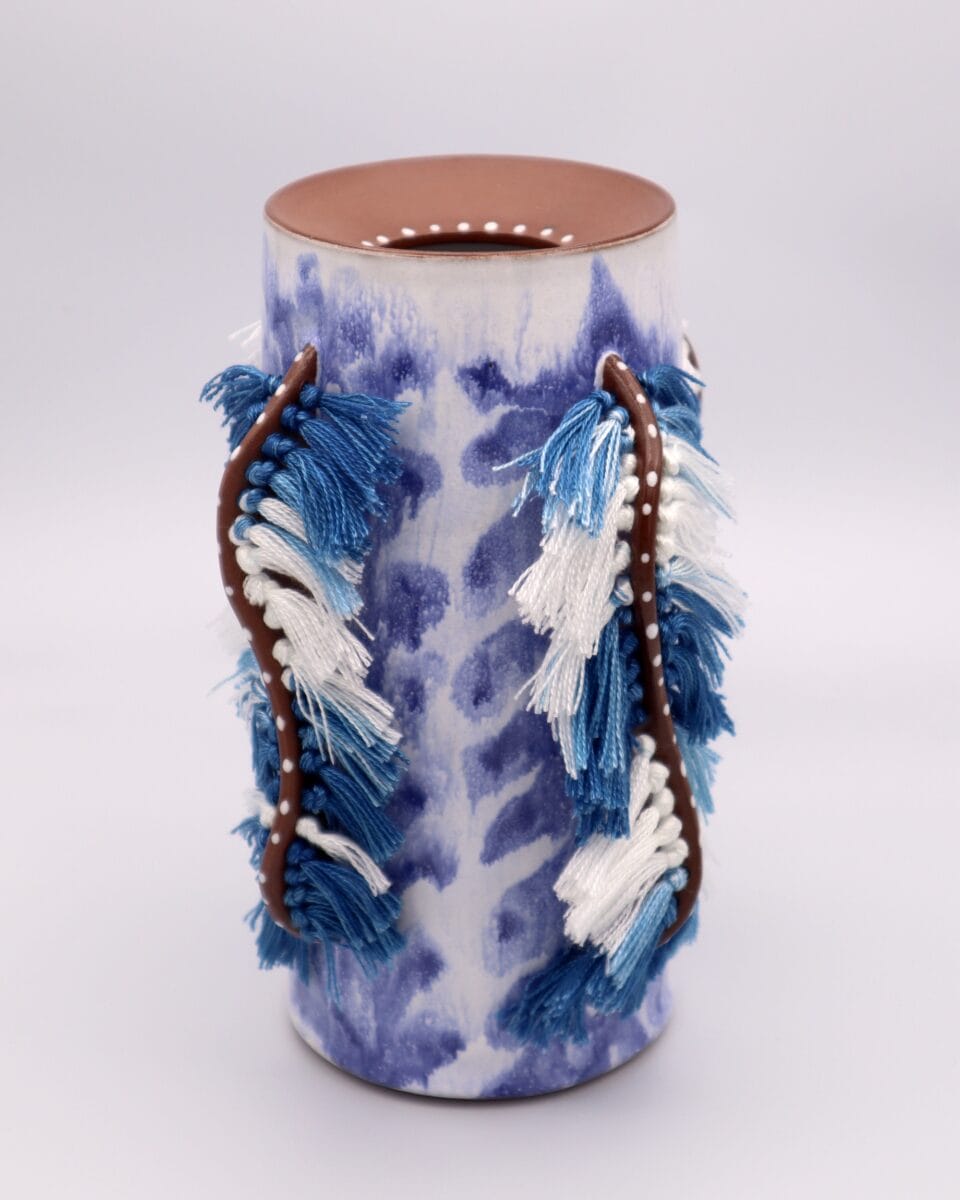 a blue-and-white glazed ceramic vessel with woven tufts of blue-and-white fiber attached vertically in places along the outside