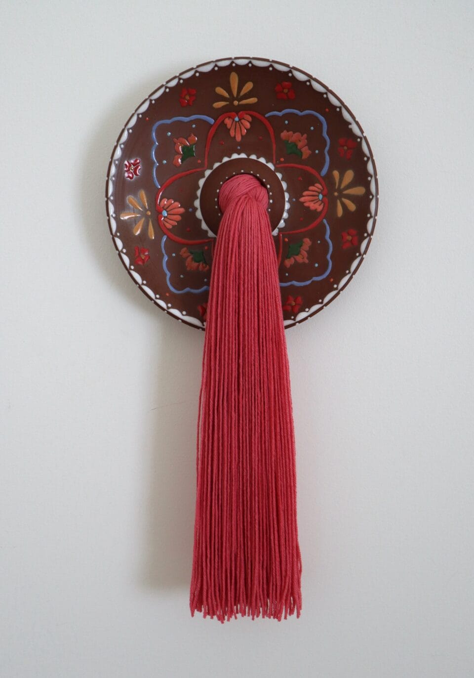 a large tuft of red fiber emerges from a central hole in a decorated ceramic plate, installed on a white wall