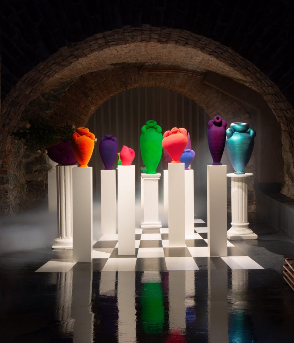 an installation view of numerous neon-colored ceramic vessels displayed on white pedestals and dramatically lit against a dark background