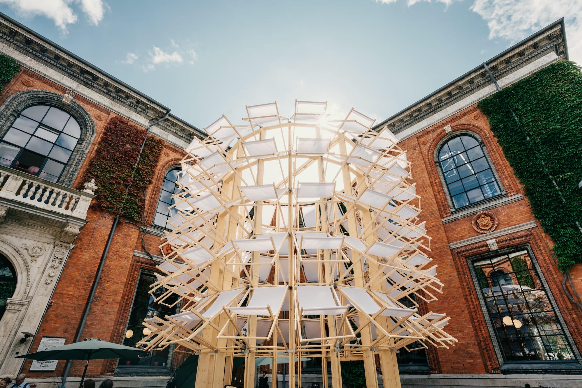 a pavilion in the courtyard of a museum made from dozens of white and timber folding deck chairs in a tall cylinder form