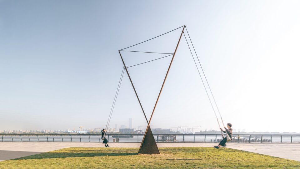 two adults swing from a massive interactive sculpture by a body of water