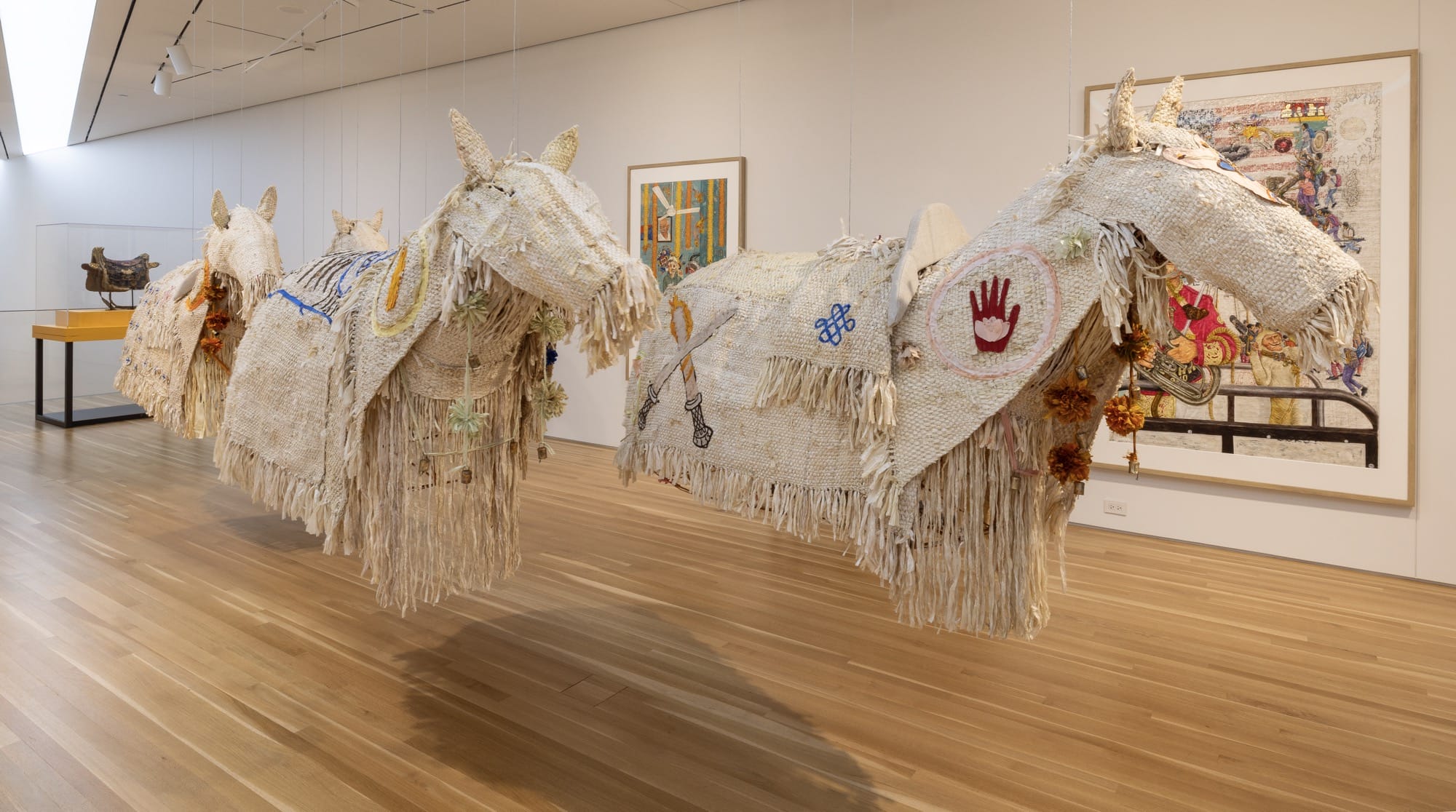 an installation of textile horses is suspended in a gallery with works on the wall and pedestal in the background