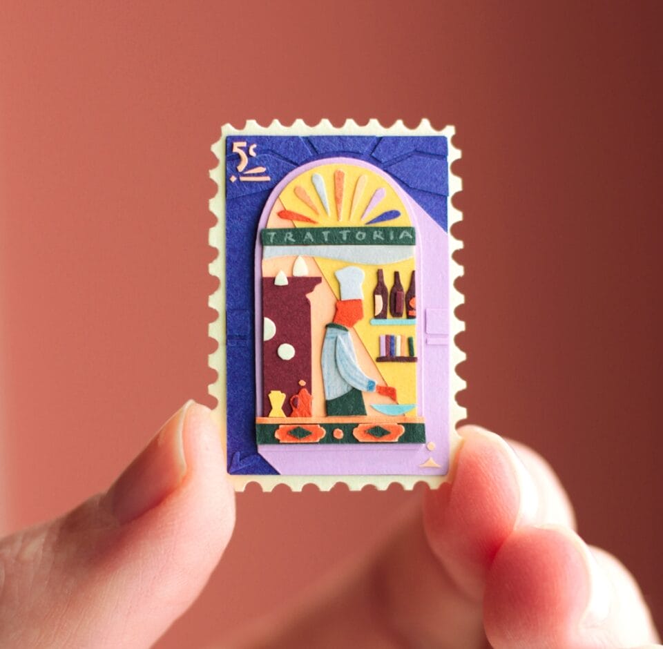 a hand holds up a small, vibrant collage in the shape of a stamp. it depicts a scene with a building and patterned designs