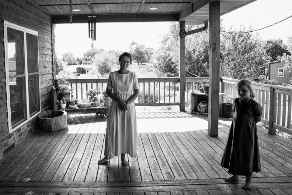 a black and white photo of a mother and daughter in long traditional dresses standing on a wood porch