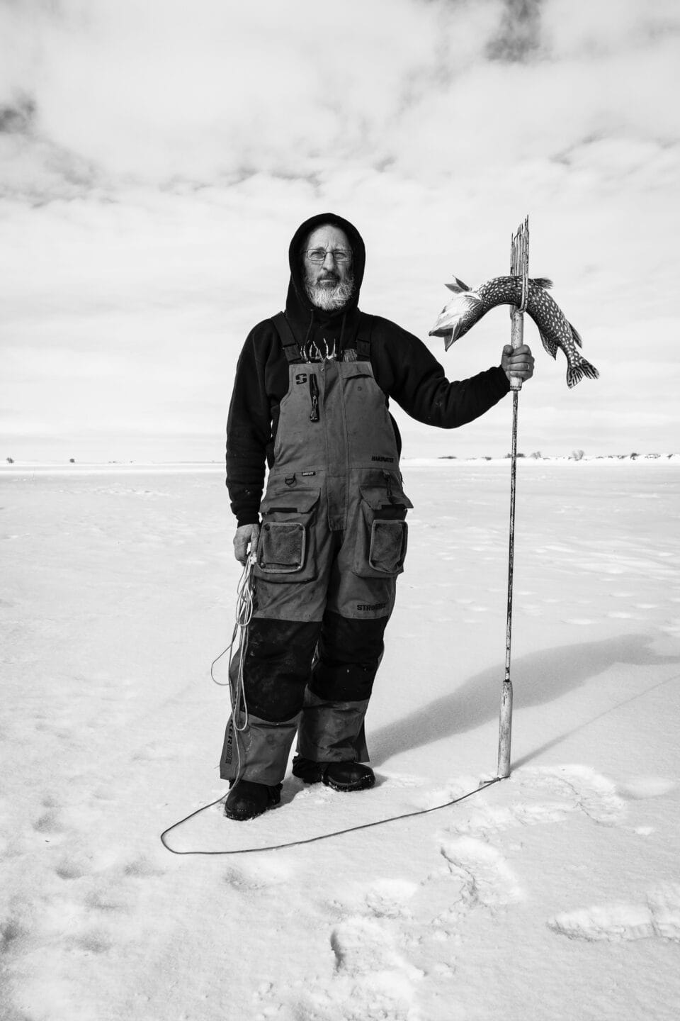 a black and white photo of a fisherman holding a speared fish on an icy body of water