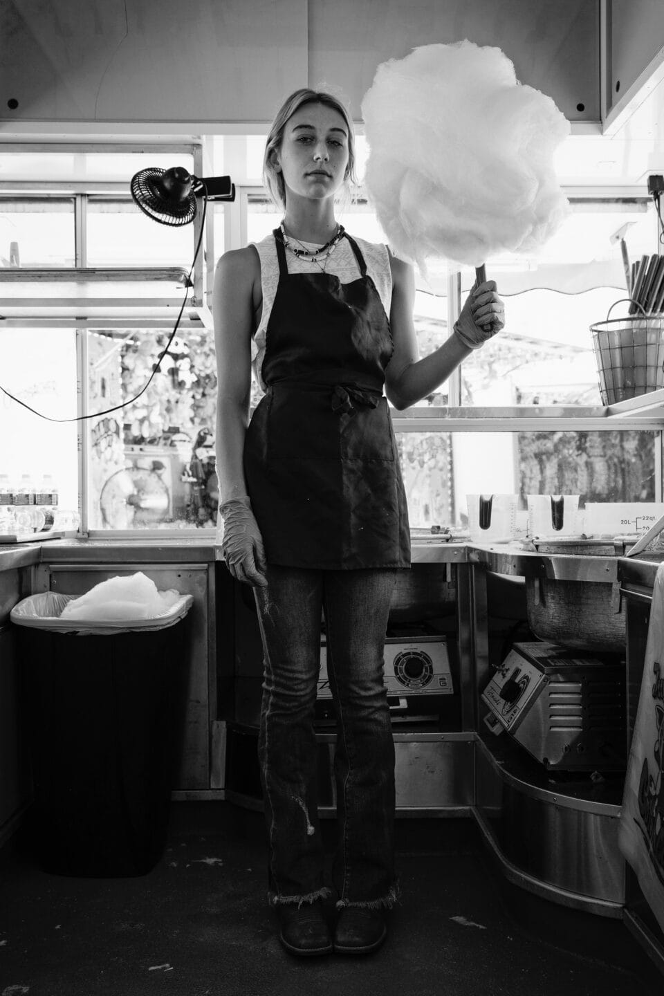 a black and white photo of a teenage girl wearing an apron holding a giant stick of cotton candy