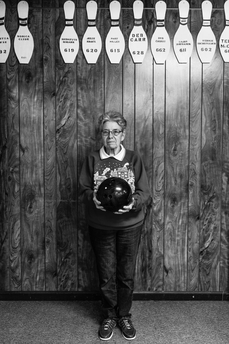 a black and white photo of an older woman posing with a black bowling ball in front of wood paneling