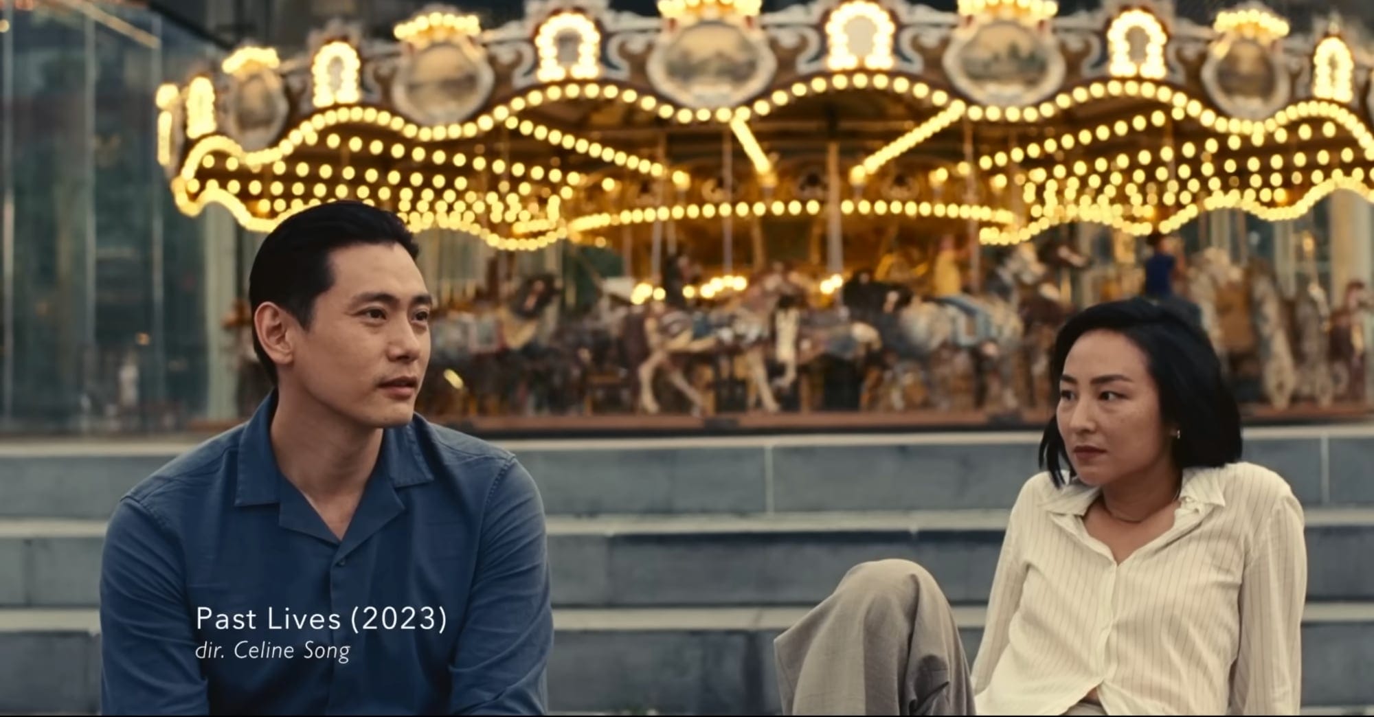 a film still from Past Lives of a man and woman sitting outside a brightly lit merry go round