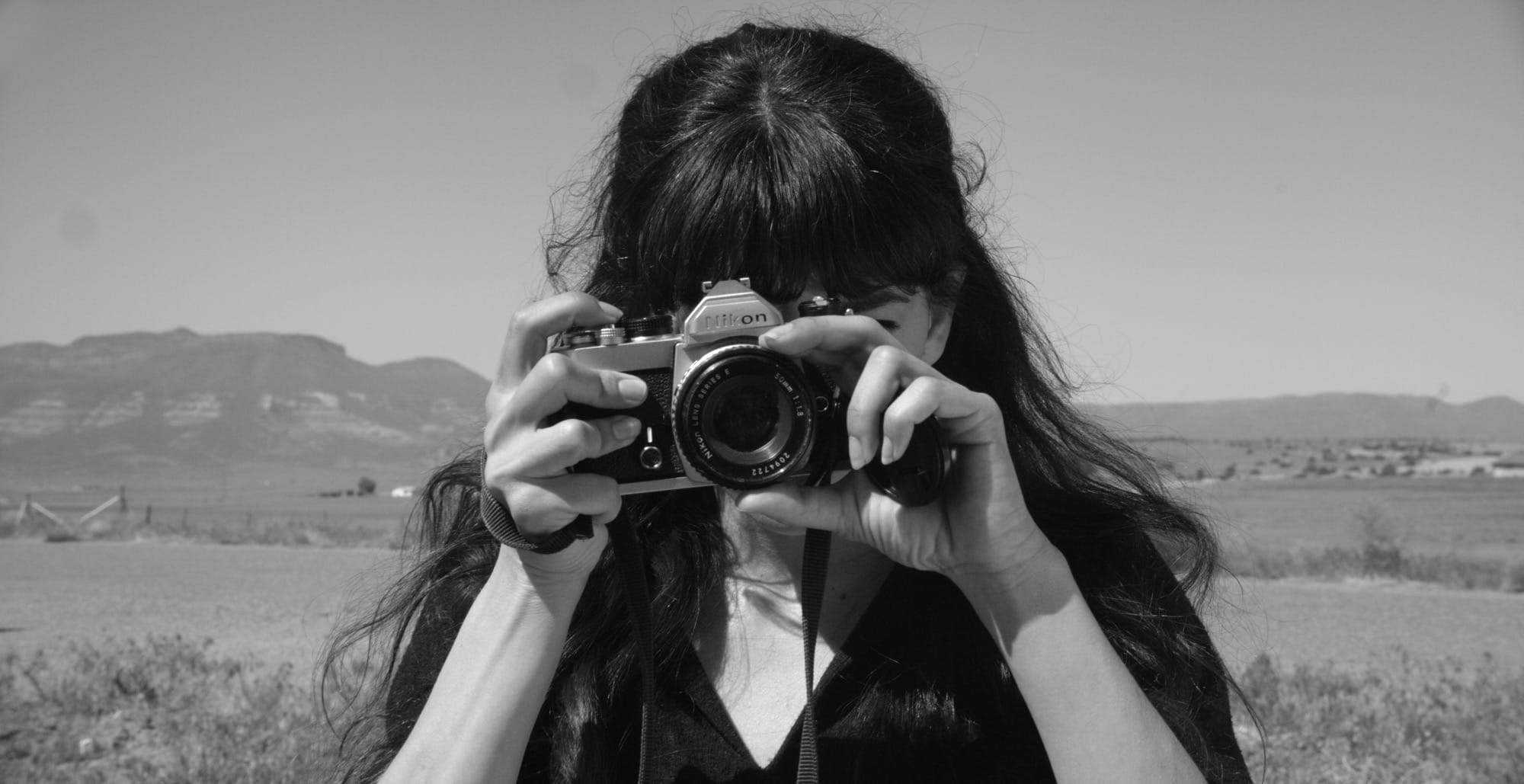 a black-and-white portrait of an anonymous woman taking a photo with an SLR camera with the desert in the background