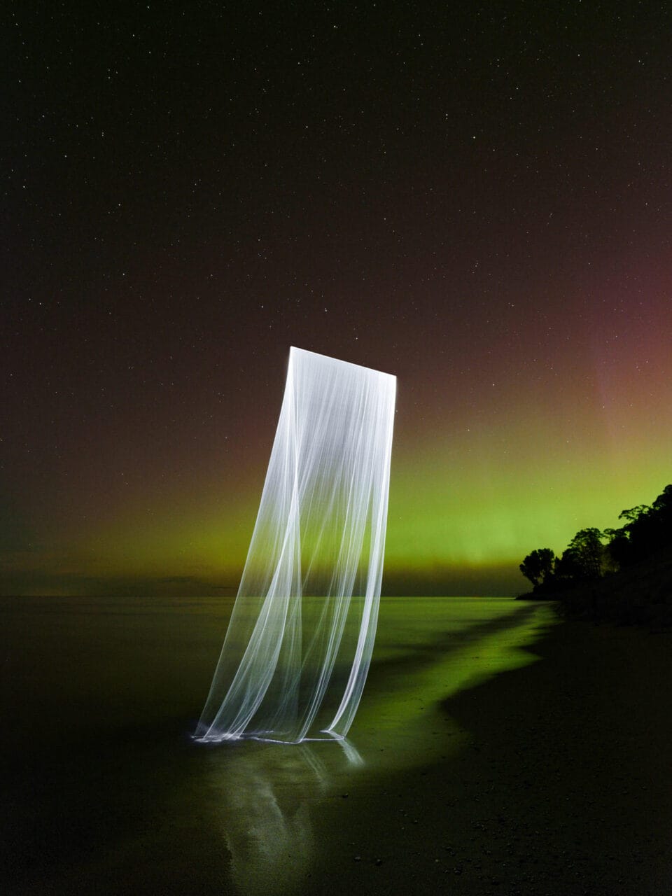 a photograph taken at night along the shore of Lake Michigan, with a green curtain of the northern lights in the distance and a laser-lit curtain of light draped over the water