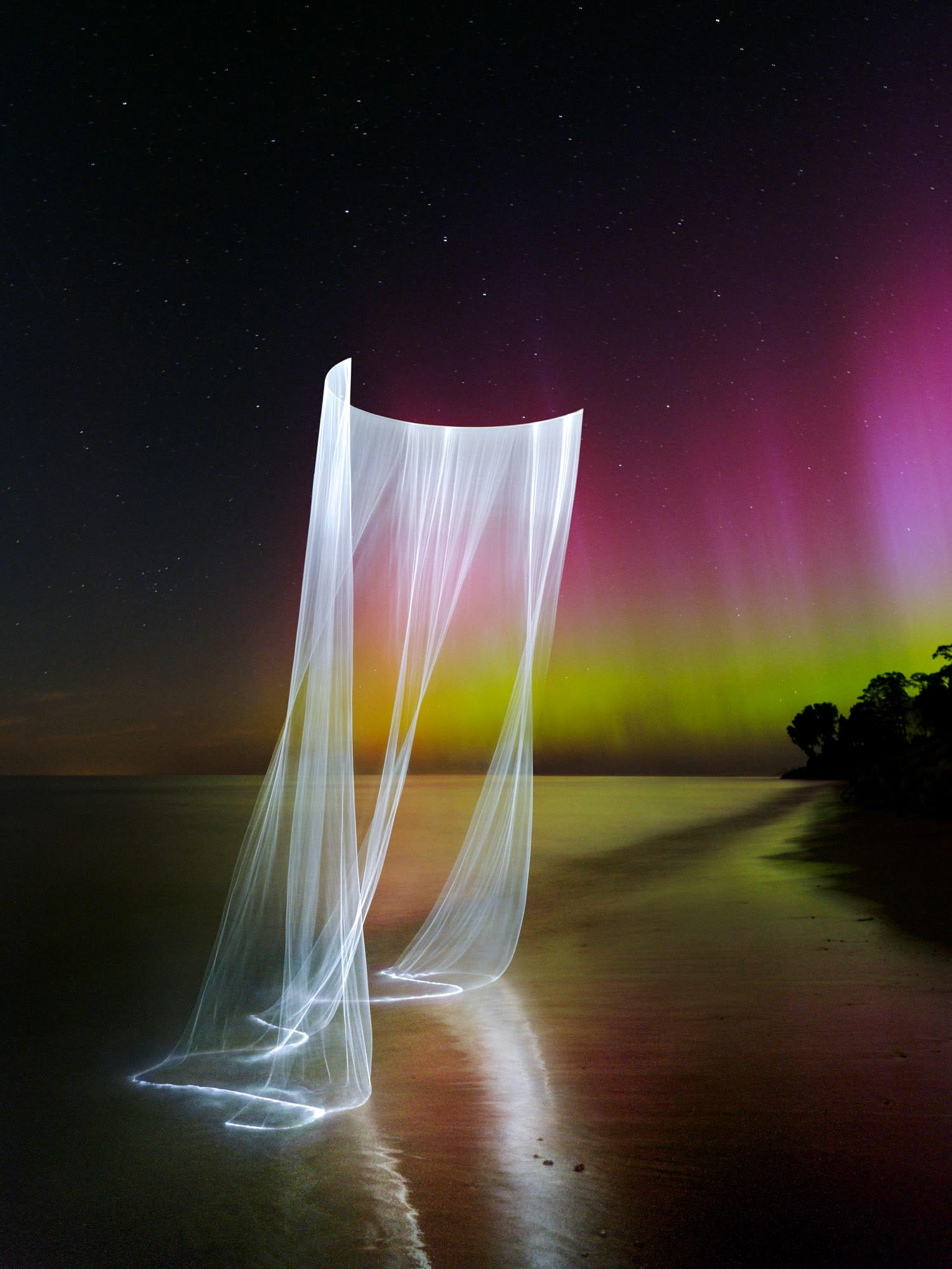 a photograph taken at night along the shore of Lake Michigan, with a magenta and green curtain of the northern lights in the distance and a laser-lit curtain of light draped over the water