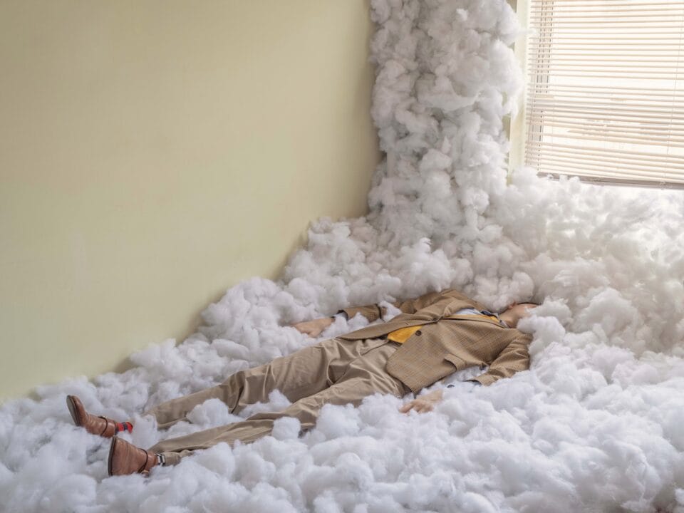 a man in a tan suit lies in puffy white batting to conceal his face